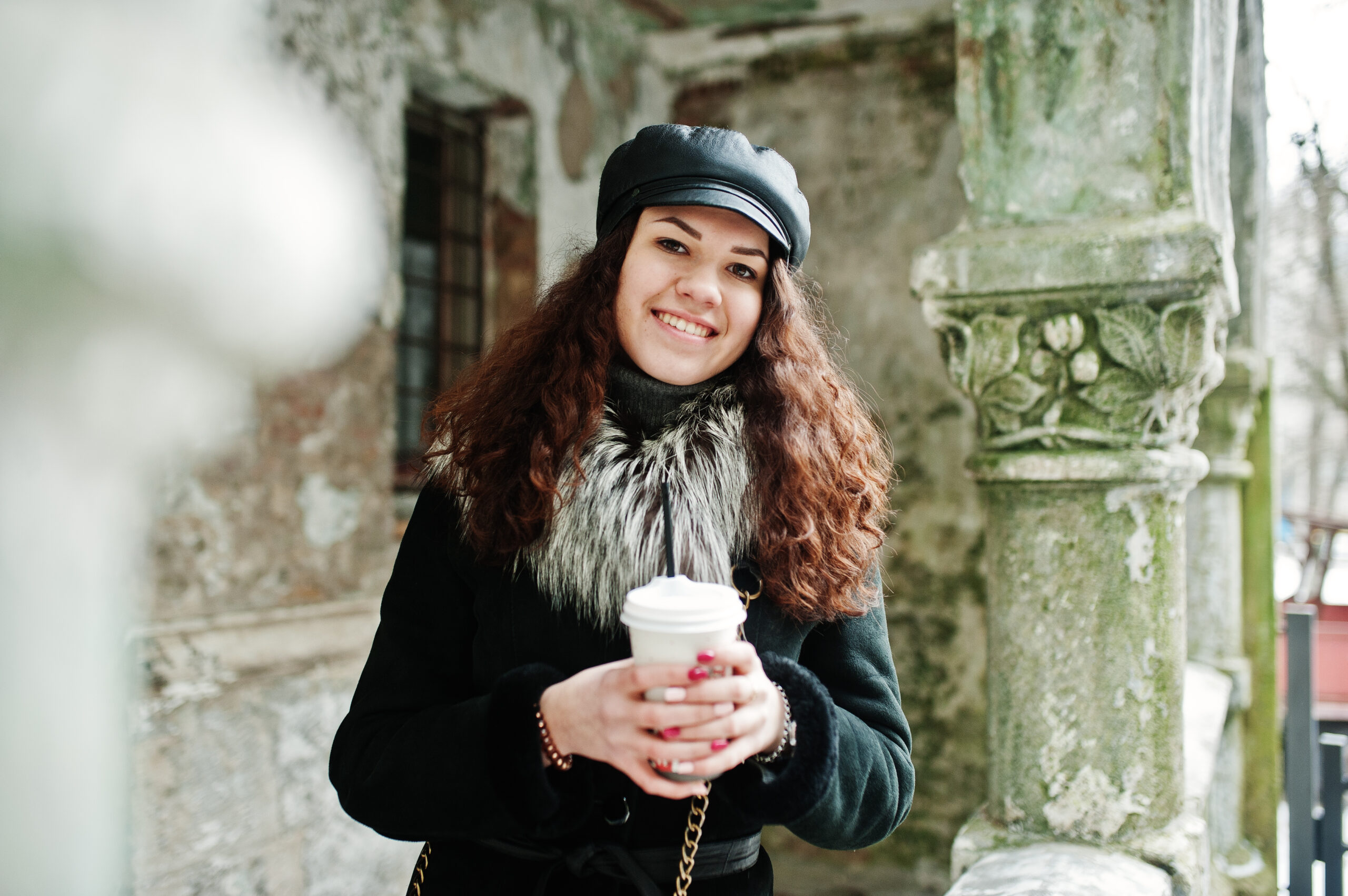 Curly mexican girl in leather cap and plastic cup of coffee at h Curly mexican girl in leather cap and plastic cup of coffee at hand walking at streets of city.